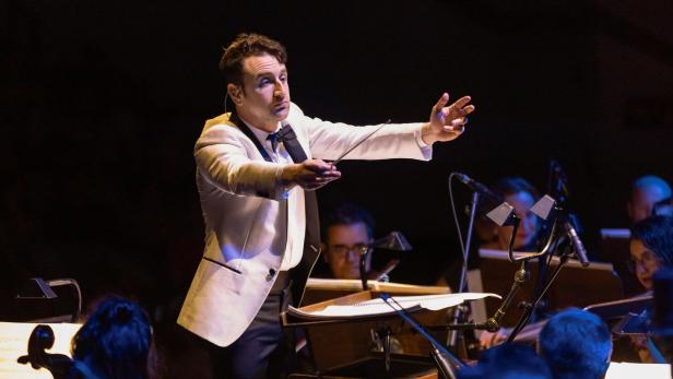Ein Dirigent in weißem Sakko leitet ein Orchester, das konzentriert auf seine Anweisungen achtet.