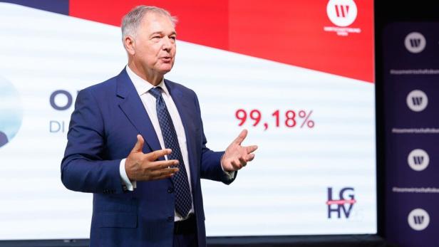 Walter Ruck  im blauen Anzug  spricht nach seiner Leinwand vor einer Leinwand, auf der  99,18 % gezeigt werden. 
