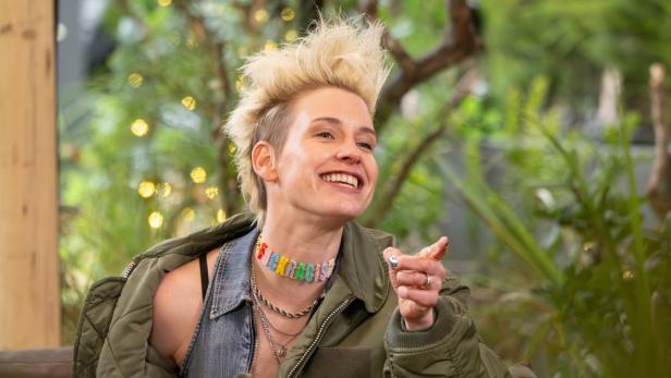 Eine lächelnde Frau mit blondem Irokesenschnitt trägt eine grüne Jacke und eine bunte Halskette.