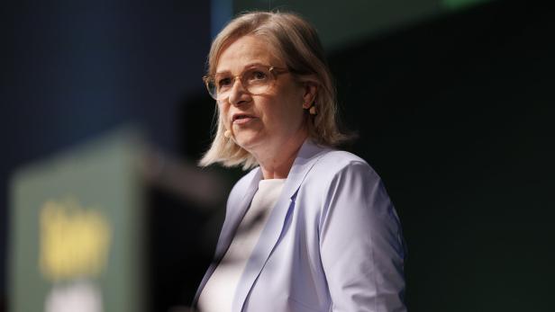 Eine Frau mit Brille und hellem Blazer spricht vor dunklem Hintergrund.