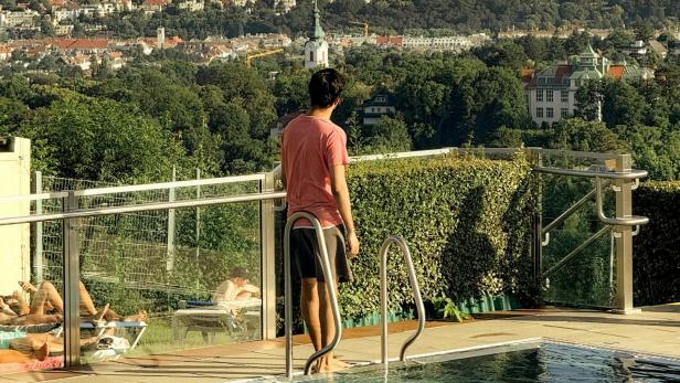 Ein junger Mann streckt sein rechtes Bein in einen Pool und blickt über die Dächer Wiens.