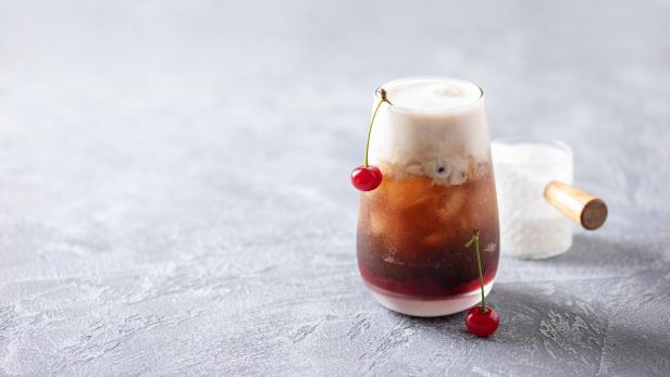 Ein Glas mit einem schaumigen Dirty Soda und Kirschen als Dekoration steht auf einem Tisch.