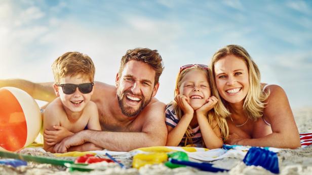 Vierköpfige Familie liegt lachend am Strand mit Strandspielzeug und Ball.