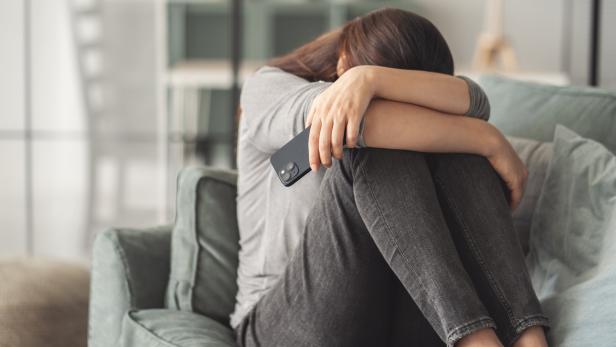 Eine Frau sitzt mit gesenktem Kopf auf einem Sofa und hält ein Smartphone in der Hand.