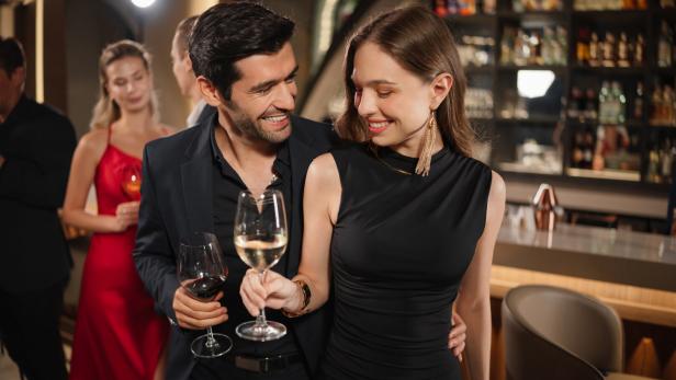 Ein elegant gekleidetes Paar steht lächelnd mit Weingläsern in der Hand an einer Bar, im Hintergrund weitere Gäste.