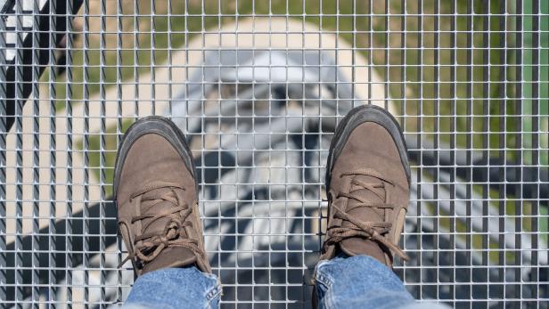 Zwei Füße in braunen Schuhen stehen auf einem Gitterrost, durch den man nach unten auf eine Treppe blickt.