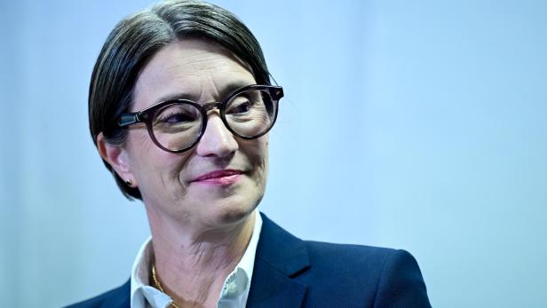 Frau mit dunklem Blazer, weißer Bluse und Brille lächelt leicht vor hellblauem Hintergrund.