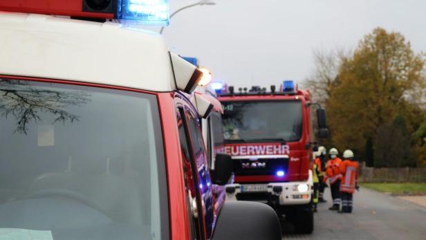 Feuerwehrautos mit Blaulicht und Einsatzkräften auf einer Straße vor herbstlichen Bäumen.
