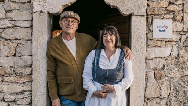 Ein kroatisches Gastgeberpaar steht lächelnd in traditioneller Kleidung vor einem Steinhaus mit dem „Local Host“-Gütesiegel an der Wand.