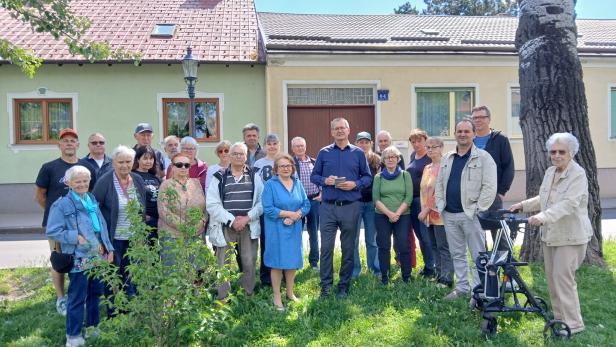 Eine große Gruppe von Menschen steht vor zwei Häusern auf einer Wiese, wobei eine ältere Frau rechts einen Rollator nutzt.