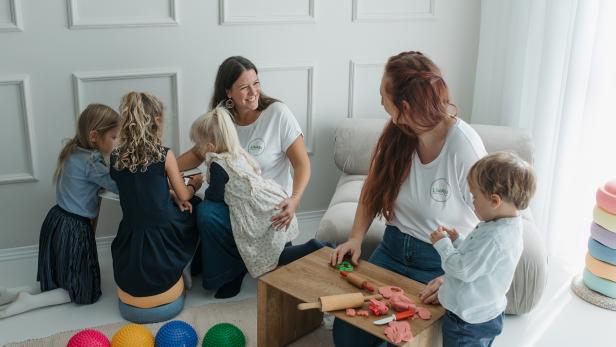 Zwei Betreuerinnen spielen und basteln mit mehreren Kindern in einem hellen Raum.