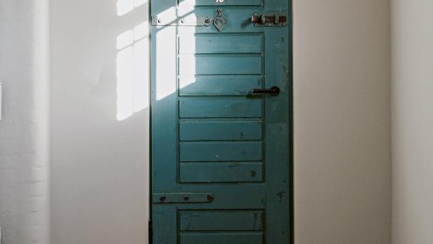 Eine alte, grüne Holztür mit der Nummer 18 in einem hellen Raum, Sonnenlicht fällt durch ein Fenster auf die Tür.