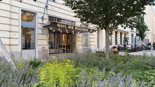 Vor einem Gebäude mit dem Schriftzug „Haus der Begegnung“ wachsen Blumen und Sträucher, daneben sitzen Menschen im Straßencafé.