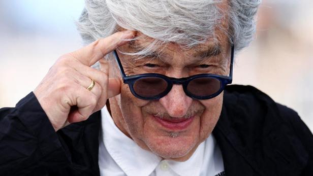 Älterer Mann mit grauen Haaren und dunkler Brille hebt zwei Finger an die Stirn und lächelt leicht.
