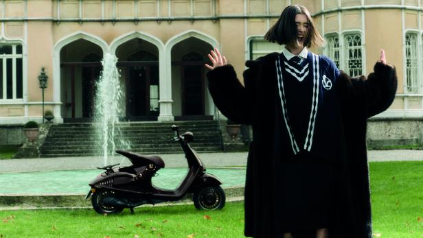 Frau mit dunklem Mantel steht vor einer Vespa und einem Brunnen vor einem historischen Gebäude.