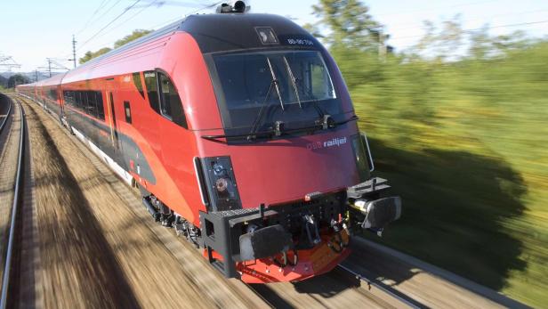 Ein roter ÖBB Railjet-Zug fährt mit hoher Geschwindigkeit durch eine grüne Landschaft.