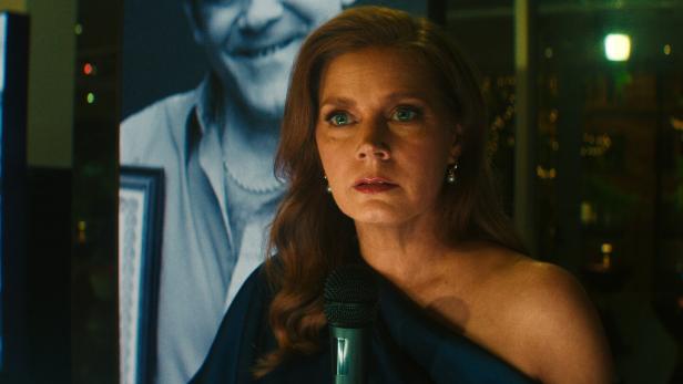 Amy Adams spricht in ein Mikrofon, während im Hintergrund ein großformatiges Schwarz-Weiß-Porträt eines Mannes zu sehen ist.