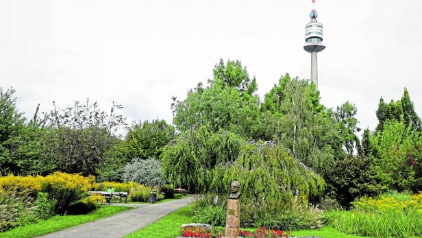 Eine Büste steht in einem grünen Park vor dem Donauturm in Wien, umgeben von Bäumen, Blumenbeeten und einem Spazierweg.