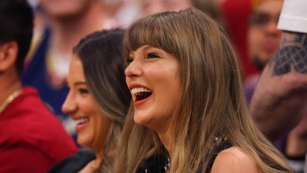 Taylor Swift lacht herzlich, während sie neben einer anderen Frau in einer Menschenmenge sitzt.