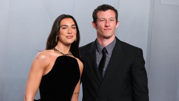 Dua Lipa und Callum Turner posieren gemeinsam vor einem hellgrauen Hintergrund.