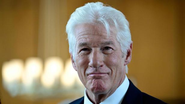 Richard Gere mit weißem Haar und einem sanften Lächeln vor einem unscharfen Hintergrund.