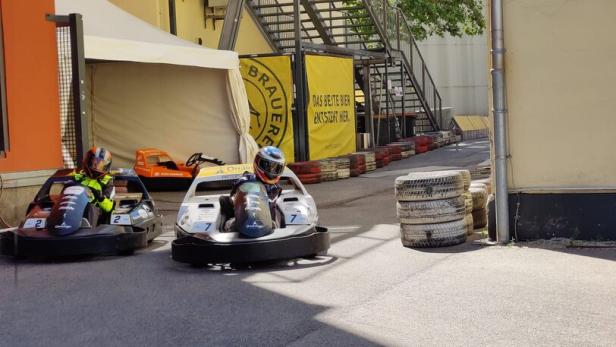 Zwei Personen fahren mit Karts auf einer Outdoor-Strecke an Gebäuden und gestapelten Reifen vorbei.