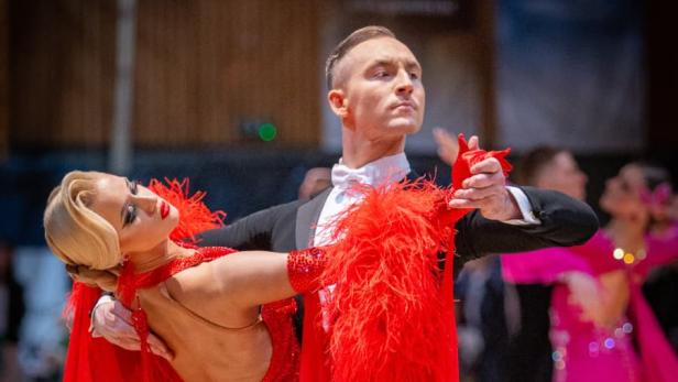Ein elegantes Tanzpaar tanzt in festlicher Kleidung, die Frau trägt ein rotes Kleid mit Federn.
