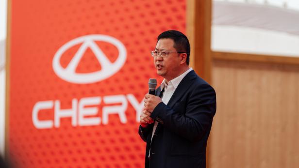Ein Mann im Anzug spricht mit Mikrofon vor einer roten Wand mit dem Chery-Logo und Schriftzug.