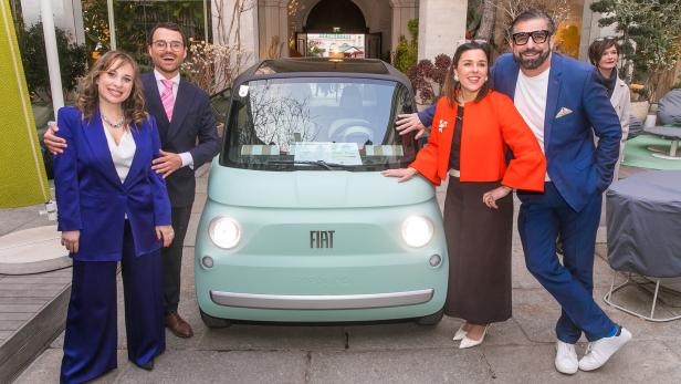 Vier elegant gekleidete Personen stehen lächelnd neben einem kleinen, mintgrünen Fiat-Auto im Freien.
