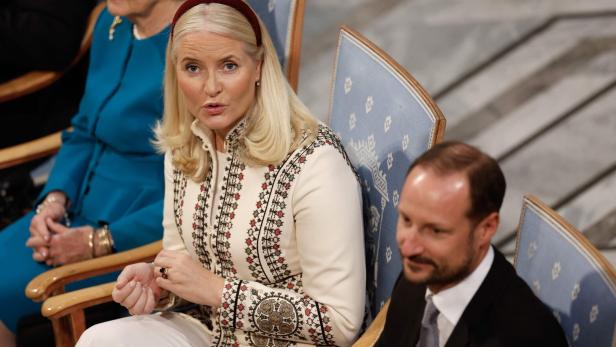 Kronprinzessin Mette-Marit von Norwegen sitzt neben Kronprinz Haakon in einem hellen, bestickten Mantel auf einem blauen Stuhl.