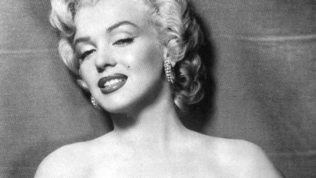 Marilyn Monroe mit blonden Locken und schulterfreiem Kleid blickt lächelnd in die Kamera.