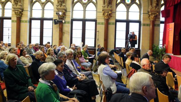 Ein Publikum sitzt in einem Saal mit hohen Fenstern und wartet auf eine Veranstaltung.