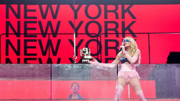 Eine Frau mit blonden Haaren und auffälligem Outfit singt auf einer Bühne vor rotem Hintergrund mit dem Schriftzug „NEW YORK“.