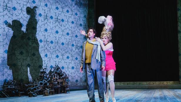 Ein Mann im Anzug und eine Frau in auffälligem pinkem Kleid stehen auf einer Theaterbühne mit gemusterter Wand und Stuhlhaufen.