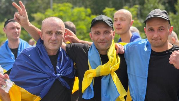 Männer, die in eine ukrainische Fahne gehüllt sind, legen sich die Arme um die Schultern und blicken in die Kamera.