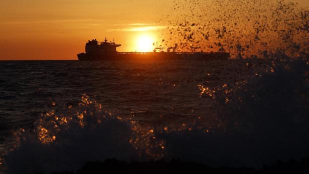 Die Silhouette eines Frachtschiffs fährt vor dem Hintergrund eines leuchtend orangefarbenen Sonnenuntergangs über das aufgewühlte Meer.