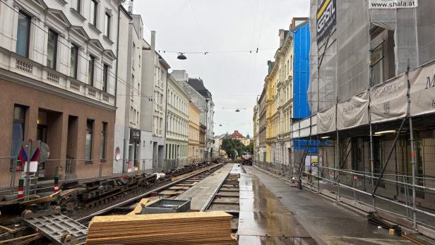 Straße mit Baustelle und offenen Straßenbahnschienen, umgeben von mehrstöckigen Häusern und Baugerüsten bei nassem Wetter.