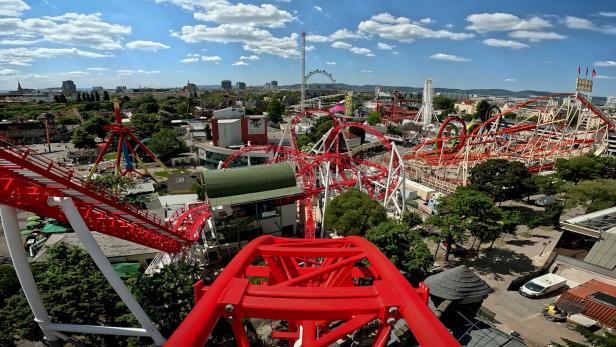 Blick von einer Achterbahn auf den Wiener Prater mit seinen Fahrgeschäften und dem Riesenrad unter einem sonnigen Himmel.