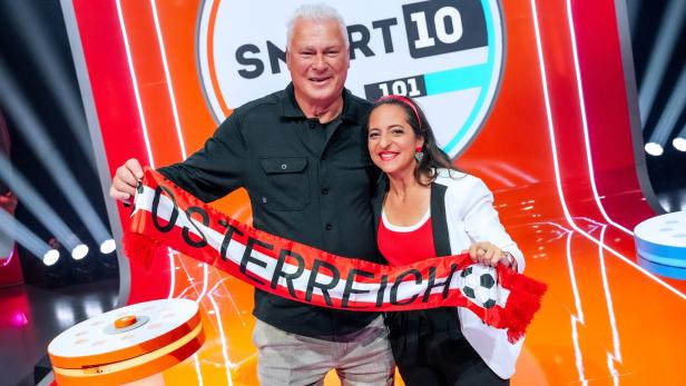 Anton Polster und Caroline Athanasiadis halten gemeinsam einen rot-weiß-roten Schal mit der Aufschrift „Österreich“ vor einer bunten Spielshow-Kulisse.
