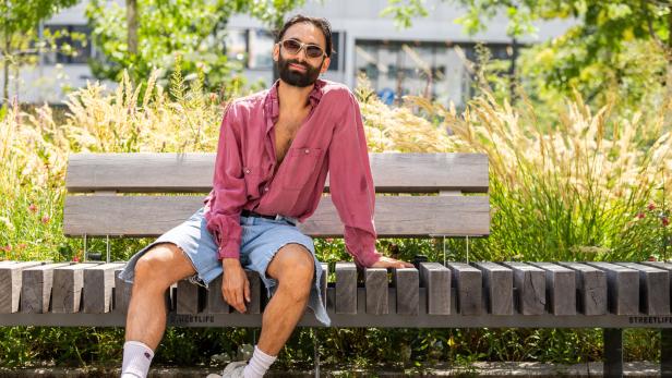 Ein Mann mit Sonnenbrille, offenem roten Hemd und Jeansshorts sitzt entspannt auf einer Holzbank im Grünen.