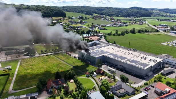 Dichter schwarzer Rauch steigt aus einem brennenden Industriegebäude in ländlicher Umgebung auf, Feuerwehr ist im Einsatz.