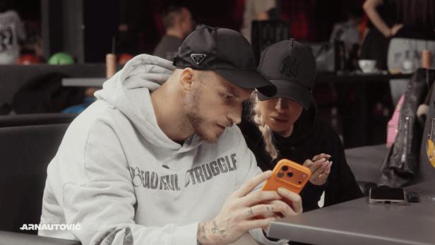 Zwei Personen mit Kappen sitzen an einem Tisch und schauen gemeinsam konzentriert auf ein orangefarbenes Smartphone.