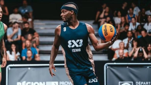 Ein Basketballspieler im blauen Trikot mit der Aufschrift „Vienna“ hält einen Ball und schaut zu einem Gegenspieler.