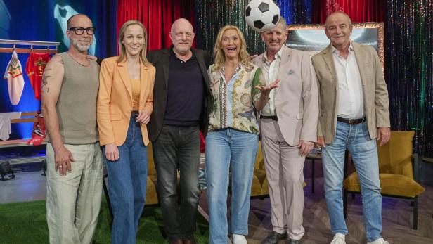 Sechs Personen posieren lachend in einem bunt dekorierten Fernsehstudio, ein Ball schwebt in der Luft.