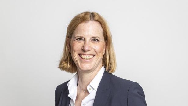 Frau mit schulterlangem blondem Haar trägt einen dunkelblauen Anzug und ein weißes Hemd und lächelt freundlich.
