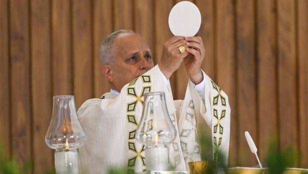 Der Papst hält während einer Messe eine große Hostie mit beiden Händen hoch, im Vordergrund stehen Kerzen.