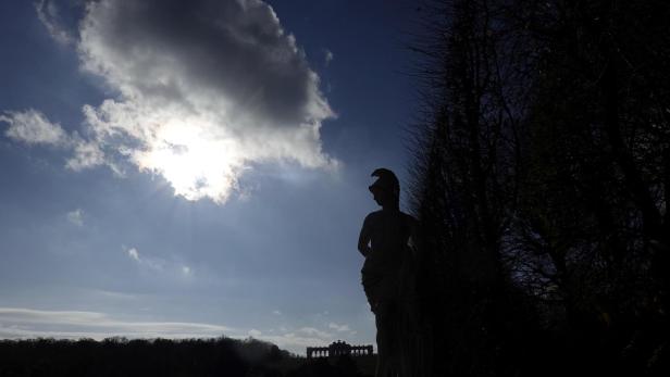Die Silhouette einer antiken Statue steht vor der Gloriette im Schlosspark Schönbrunn unter einem bewölkten Himmel.