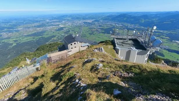 Gebäude mit Antennen auf einem bewachsenen Bergrücken, im Hintergrund weite grüne Landschaft und Berge unter blauem Himmel.