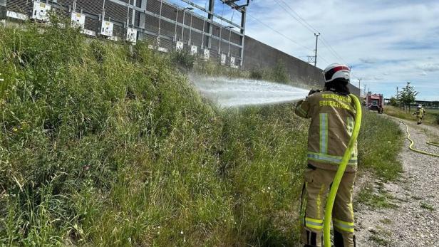 Ein Feuerwehrmann löscht mit einem Schlauch brennendes Gras neben einer Stromanlage unter freiem Himmel.