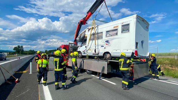 Mehrere Feuerwehrleute bergen mit einem Kran ein beschädigtes Wohnmobil auf einer Autobahn.